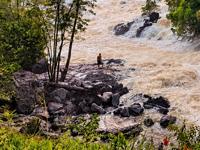 A visitor stands on dark granite rocks at the edge of the rapids, dwarfed by the falls.